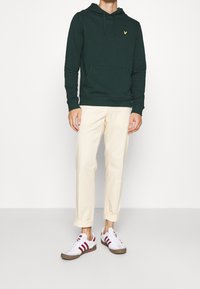 Felpa in cotone verde scuro con tasca frontale, abbinata a pantaloni color crema e sneakers bianche con strisce rosse e suola marrone.