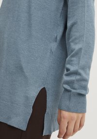 Pull en tricot bleu clair avec une fente sur le côté, des poignets côtelés et une coupe décontractée, présenté sur un fond uni.