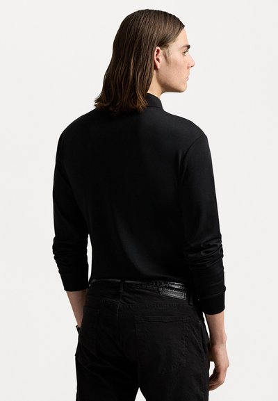 Black long-sleeve top with a high collar and relaxed fit. Smooth fabric, ribbed cuffs, viewed from the back, paired with black pants.