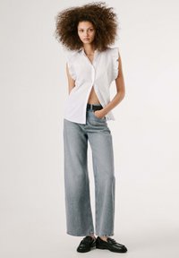 White sleeveless blouse with ruffled shoulders paired with light blue wide-leg jeans and black loafers. Plain background, minimal design.