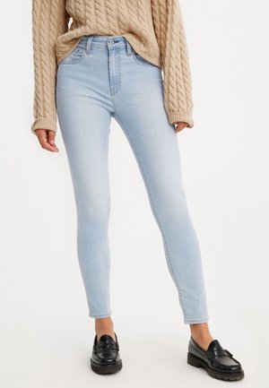 Femme portant un jean skinny bleu clair, un pull beige à torsades et des mocassins en cuir noirs, debout devant un fond blanc.