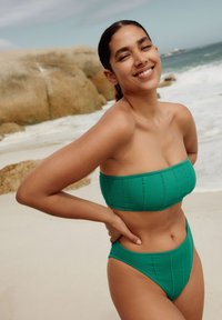 Zelená bikiny souprava s žebrovanou texturou, obsahující bandeau top s vertikálními švy a kalhotky s vysokým pasem. Hladká textura na pozadí pláže.