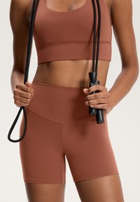 Rostfärgad sportbehå och högmidjade shorts i stretchigt material, håller en svart hopprep, visar en slät textur och en figursydd design.