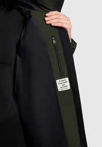 Zwarte jas met een groene voering, voorzien van een zak met rits en een label met tekst. Soepele textuur, ontworpen voor warmte en comfort.