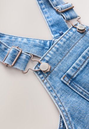 Salopette en denim bleu avec bretelles réglables, ferrures métalliques et une poche avant. Présente des coutures visibles et une finition en tissu texturé.