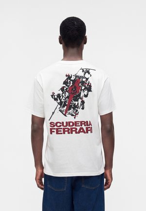 Personne portant un T-shirt blanc avec un graphisme rouge et noir d'une voiture de course et le texte "Scuderia Ferrari" au dos, associé à un jean bleu.