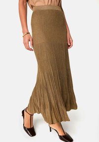Jupe maxi plissée dorée-brune métallisée avec une taille ajustée, une forme fluide et une texture douce. Associée à des chaussures à talons sombres.