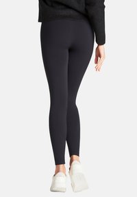 Leggings noirs en tissu extensible à texture lisse ; coupe ajustée s'étendant jusqu'aux chevilles, assortis à des chaussures blanches.