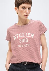 Άτομο με κοντά μαλλιά που φοράει κόκκινη και άσπρη ριγέ μπλούζα με το κείμενο "ATELIER 2010 MOS MOSH" και σκούρα παντελόνια με ζώνη, ποζάροντας μπροστά σε μονόχρωμο φόντο.