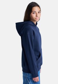 Haut à capuche bleu marine en mélange de coton avec une poche frontale, des poignets côtelés et une capuche à cordon, associé à un jean en denim bleu.
