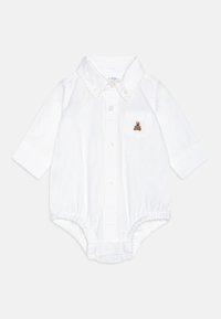 Balta medvilninė kūdikio jumpsuit su ilgomis rankovėmis, mygtukų uždengimu priekyje ir maža krūtinės kišene, kurioje išsiuvinėtas rudas lokys. Minkšta tekstūra.