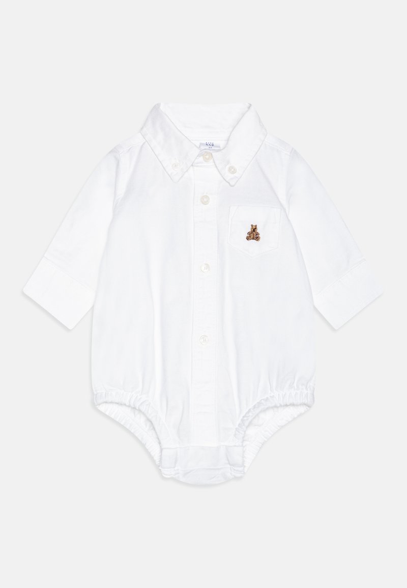 Balta medvilninė kūdikio jumpsuit su ilgomis rankovėmis, mygtukų uždengimu priekyje ir maža krūtinės kišene, kurioje išsiuvinėtas rudas lokys. Minkšta tekstūra.