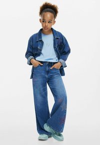 Jeansjacka med stjärndetaljer, blå T-shirt, vida jeans med stjärnmönster och turkosa sneakers. Enfaldig design, avslappnad passform.