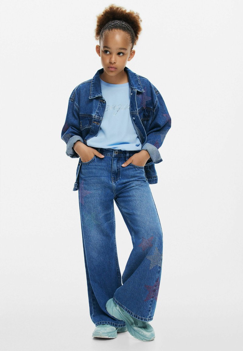 Jeansjacka med stjärndetaljer, blå T-shirt, vida jeans med stjärnmönster och turkosa sneakers. Enfaldig design, avslappnad passform.