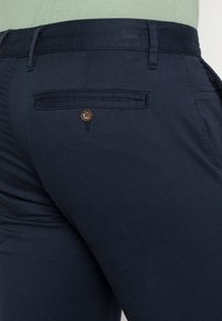 Pantalons bleu marine avec une texture lisse, une coupe ajustée et une poche arrière avec un détail de bouton marron.