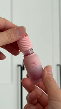 Bottiglia di vetro con finitura rosa satinata, trasparente al centro, chiusa con un tappo rosa rigato. Il testo sulla bottiglia recita "INK AIRY VELVET 21."