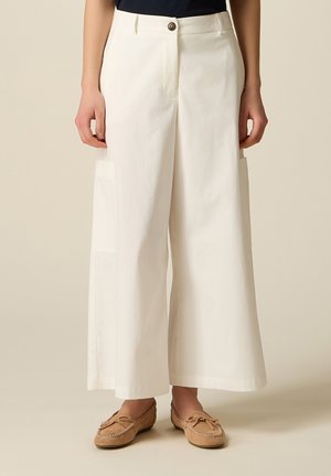 Donna che indossa pantaloni bianchi a gamba larga e vita alta con tasche laterali e mocassini color cuoio, in piedi davanti a uno sfondo semplice.