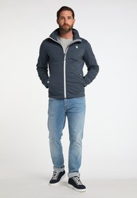 Chaqueta softshell en navy oscuro con cremallera blanca, cuello alzado y dos bolsillos frontales. Combinada con jeans azul claro y zapatillas oscuras.