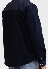 Man in een donkerblauwe corduroy shirt en lichte spijkerbroek, gezien vanaf de achterkant, tegen een vlakke lichte achtergrond.