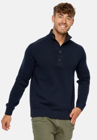 Mørkeblå strikket sweater med høj krave, knaplukning og ribbede manchetter og kant. Bæres over olivengrønne bukser.