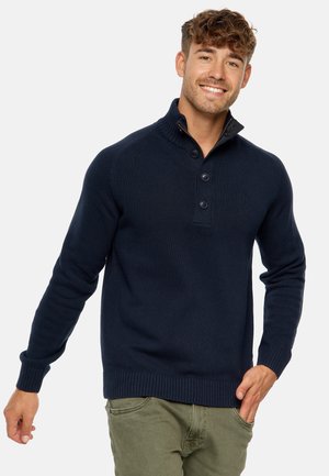 Suéter de punto azul marino con cuello alto, tapeta de botones y puños y dobladillo acanalados. Lleva puesto encima de pantalones verde olivo.