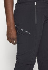 Gros plan d'une personne portant un pantalon de plein air noir Vaude avec des poches zippées sur la cuisse et près de la taille.