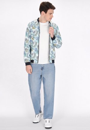Lichte bomberjack met een loofpatroon in blauw en geel, zwarte geribbelde manchetten, gecombineerd met lichtblauwe jeans en witte sneakers.