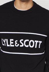 Pull en maille noire avec l'inscription LYLE & SCOTT en lettres blanches audacieuses, encadrée par une ligne blanche horizontale traversant la poitrine.