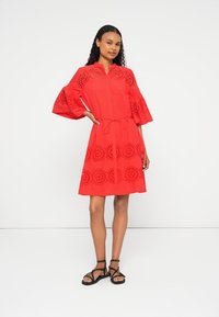 Vestido vermelho de manga curta com padrão de recortes florais, decote redondo, cintura ajustada e saia evasê. Usado com sandálias pretas de tiras.
