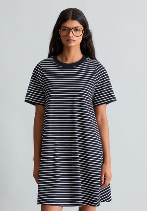 Femme aux longs cheveux foncés portant de grandes lunettes et une robe à manches courtes rayée horizontalement noir et blanc, debout sur un fond clair uni.