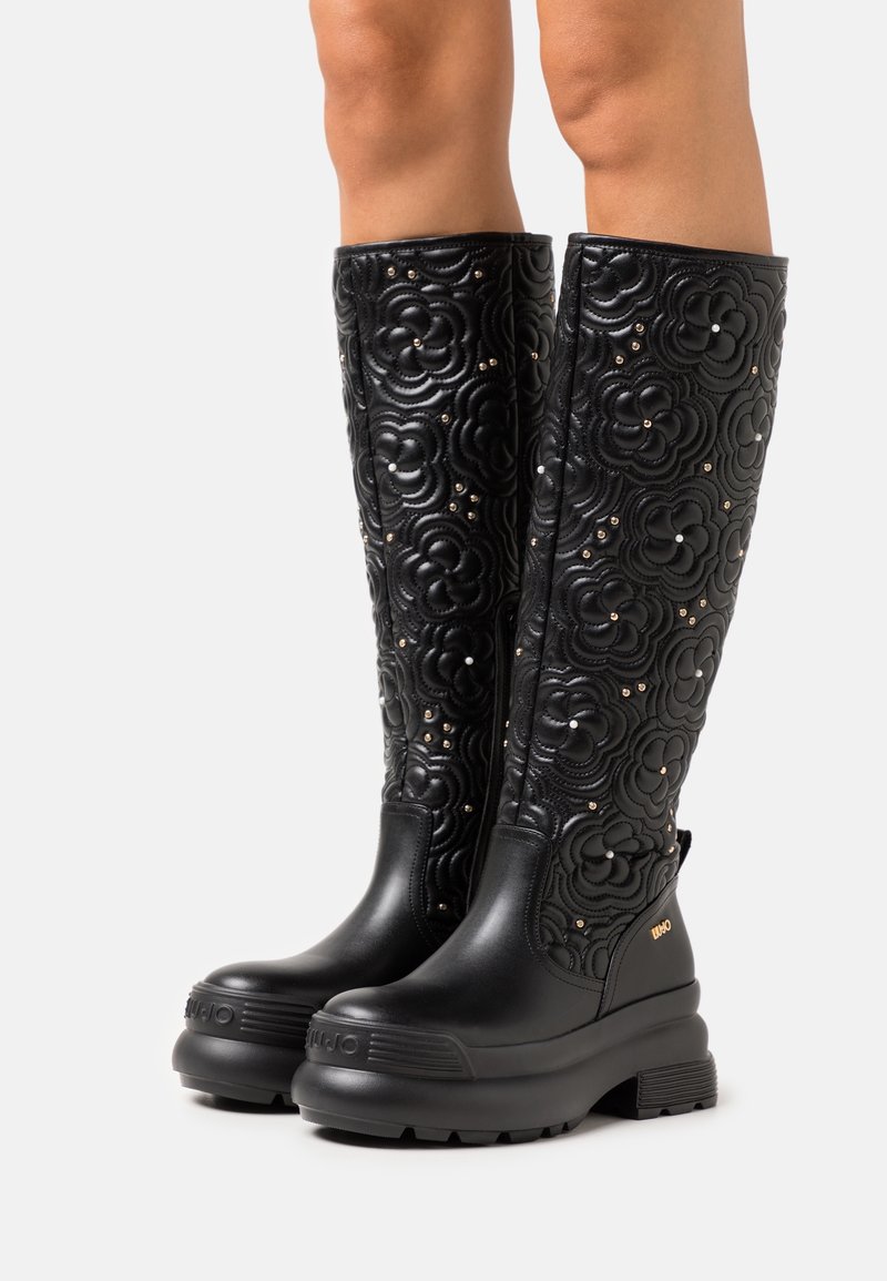 Bottes noires montantes jusqu'aux genoux avec motifs matelassés floraux et petites clous, portées par une personne aux jambes nues, avec des semelles épaisses compensées.