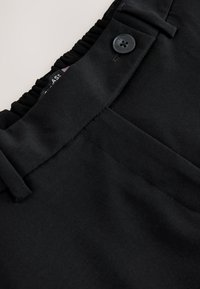 Gros plan sur la ceinture d'un pantalon noir avec dos élastique, passants de ceinture et fermeture par bouton, sur fond blanc.