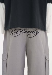 Graue Cargo-Hosen mit dem gestickten Markennamen "Ed Hardy" in großer schwarzer Schrift auf der Rückseite, kombiniert mit einem schwarzen T-Shirt.