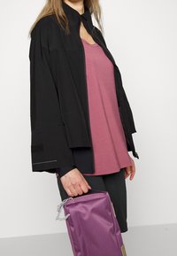 Czarna kurtka na zamek z szerokimi rękawami, w połączeniu z różowym topem z szerokim dekoltem. Trzyma fioletową prostokątną torbę z zamkiem błyskawicznym.