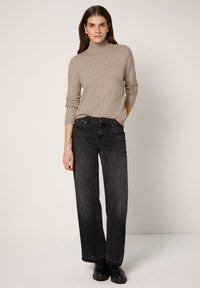 Khaki Rollkragenpullover mit gerippten Bündchen, kombiniert mit schwarzen Jeans mit weitem Bein. Das Modell steht vor einem neutralen Hintergrund und trägt schwarze Stiefel.