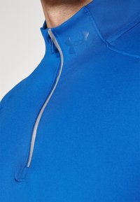 Lähellä oleva kuva henkilöstä, joka pitää yllään sinistä neljännekseltä vetoketjullista urheilupaitaa, jossa on heijastinraitana vetoketjun reunassa ja vaalea Under Armour -logo rinnassa.