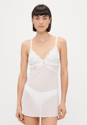 Witte kant chemise met dunne verstelbare bandjes, voorzien van een gegolfde rand, semi-doorzichtige stof en een delicaat bloemenpatroon.