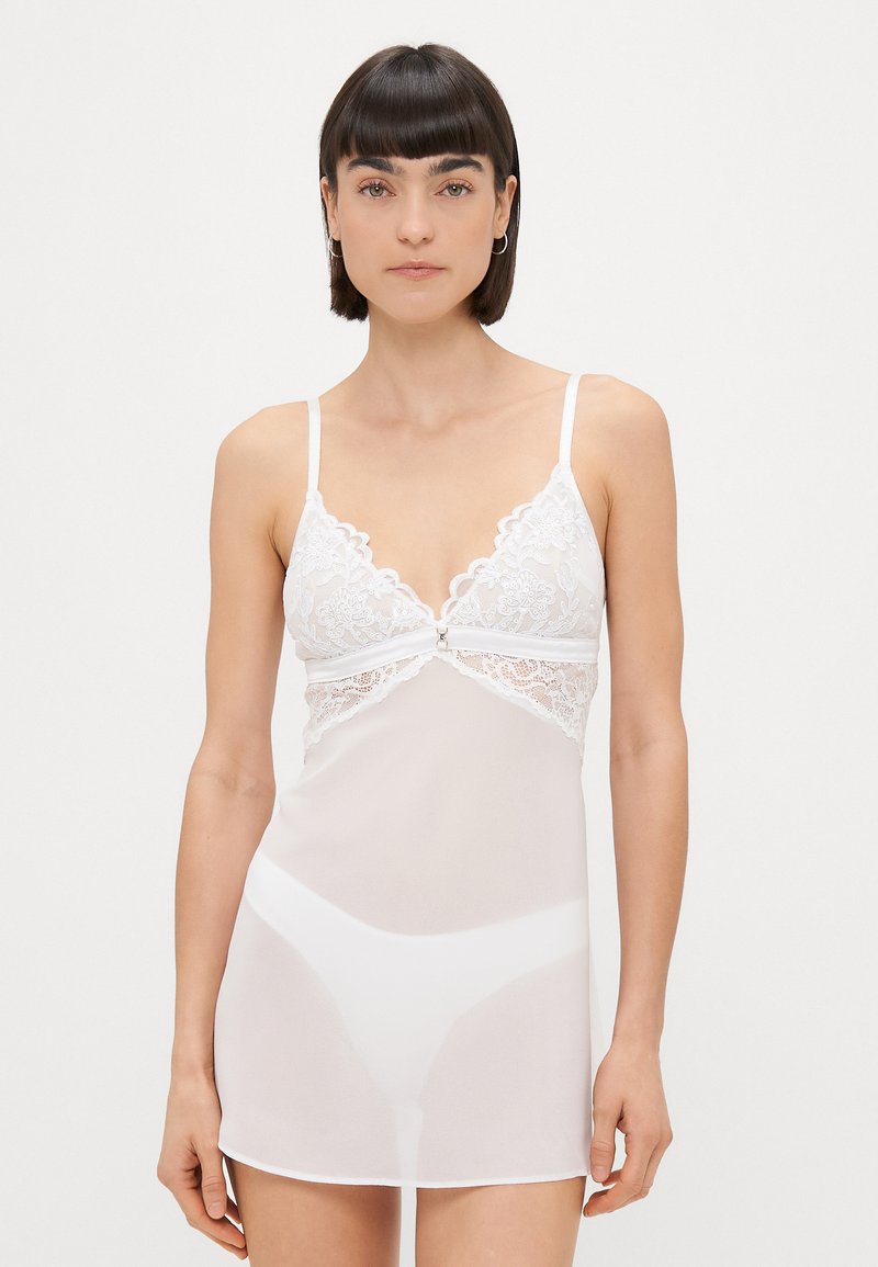 Cămașă albă din dantelă cu bretele subțiri ajustabile, având o margine ondulată, material semi-transparent și un model floral delicat.