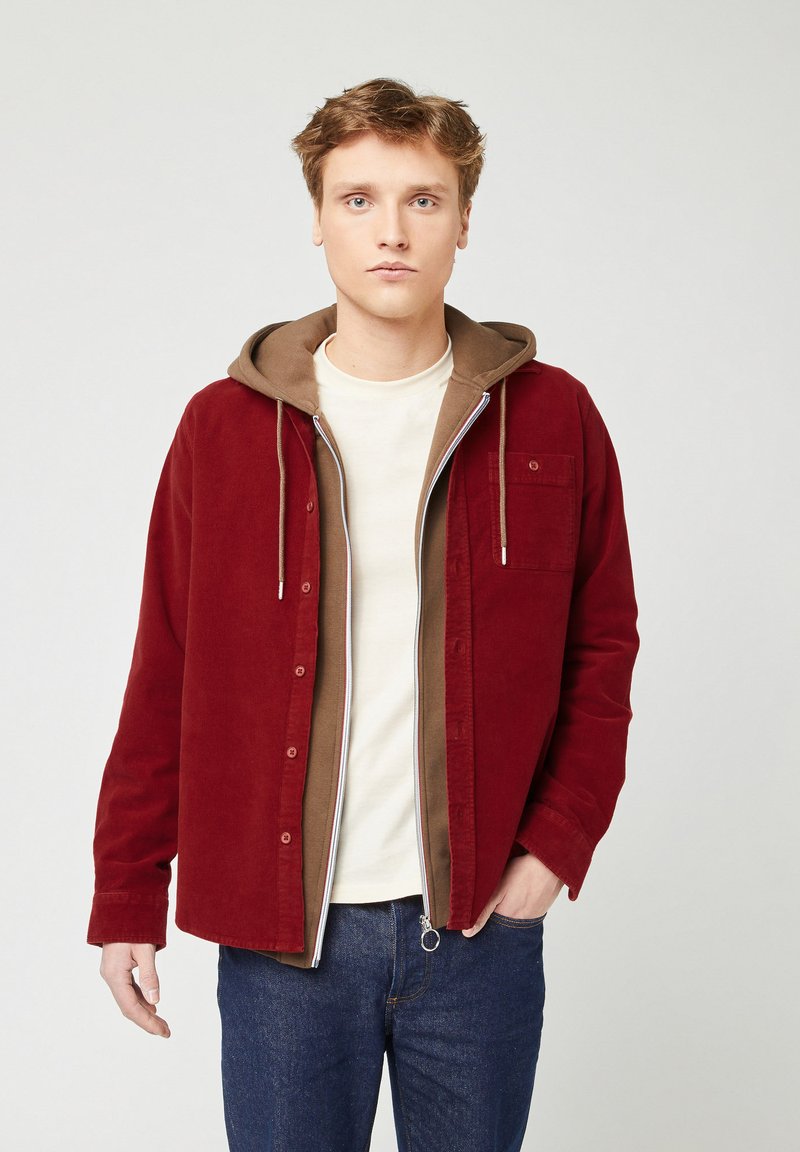 Giovane uomo che indossa abiti casual a strati: t-shirt beige, felpa marrone con zip e giacca rossa aperta, in piedi su uno sfondo semplice.