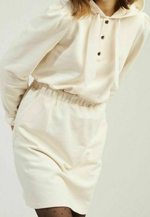 Kobieta ubrana w kremową, długorękawową sukienkę z kapturem, z czarnymi guzikami i marszczonym taliowaniem, do której dobrała czarne przezroczyste rajstopy w kropki.