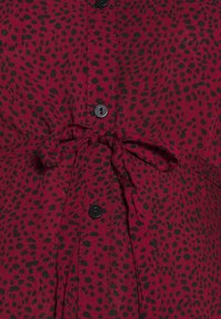 Bordeauxfarbenes Hemd mit schwarzem Tupfenmuster, verfügt über einen gebundenen Taillendetail und schwarze Knöpfe entlang der Vorderseite.