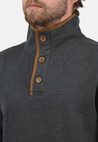Charcoal gray pullover with a high collar, featuring a brown trim and three wooden buttons on the placket. Soft, textured fabric.