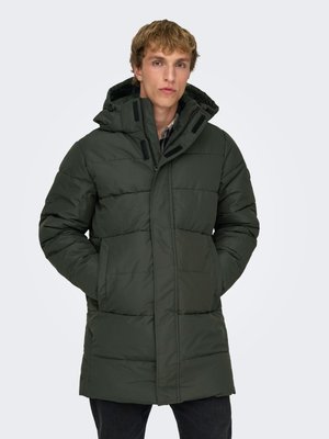 Young man wearing a dark green padded winter coat with a hood, hands in pockets, standing against a plain light background.