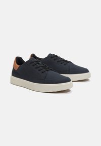 Marineblauwe canvas sneakers met een tan leren hielaccent. Voorzien van een ronde neus, vetersluiting aan de voorkant en een crèmekleurige rubbersole.