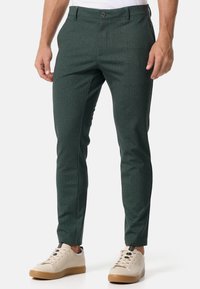 Grüne Hosen im Fischgrätmuster mit slim-fit, ausgestattet mit einem Vorderknopfverschluss, Gürtelschlaufen und Stofftextur. Kombiniert mit hellen Sneakers.