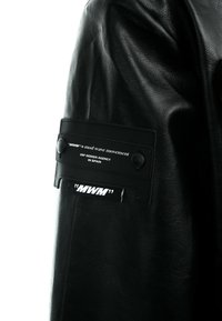 Chaqueta de cuero negra con una textura suave, con un parche prominente en la manga que muestra el logo de la marca y detalles en texto.