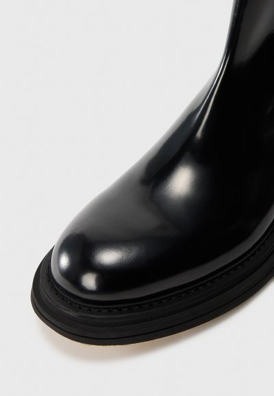 Botín de cuero negro pulido hasta el tobillo con punta redonda, que presenta una suela gruesa negra y una superficie superior elegante y suave.