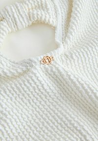 Baltas tekstūruotas drabužis su smock siuvimu, apvalia iškirpte ir auksinio tono apvalia sagtele užsegimu. Lygus, lengvas audinys.