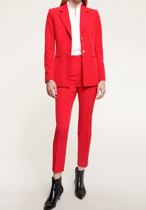 Costume rouge avec une veste ajustée et un pantalon sur mesure. La veste a une fermeture à un seul bouton et des revers en dents de poisson. Porté par-dessus un chemisier blanc.