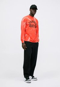 Maglietta a maniche lunghe rossa con effetto tie-dye e scritta nera "swamp queen", abbinata a pantaloni neri a taglio largo e sneakers bianche e nere.